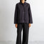 A woman stands in a studio wearing a dark charcoal button-front jacket with front patch pockets, black wide-leg pants, and black shoes against a plain white wall.