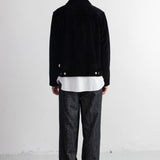 A man viewed from behind wearing a black jacket over a white shirt, paired with dark gray textured trousers and black shoes, standing against a plain white wall.