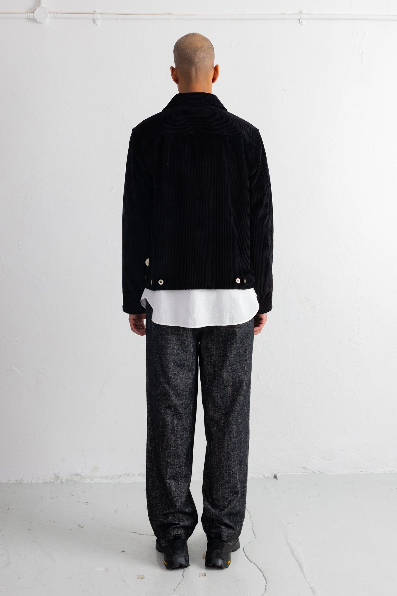 A man viewed from behind wearing a black jacket over a white shirt, paired with dark gray textured trousers and black shoes, standing against a plain white wall.