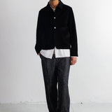 A man stands in a minimalist studio wearing a black corduroy jacket over a white shirt, paired with dark gray textured trousers and black shoes.