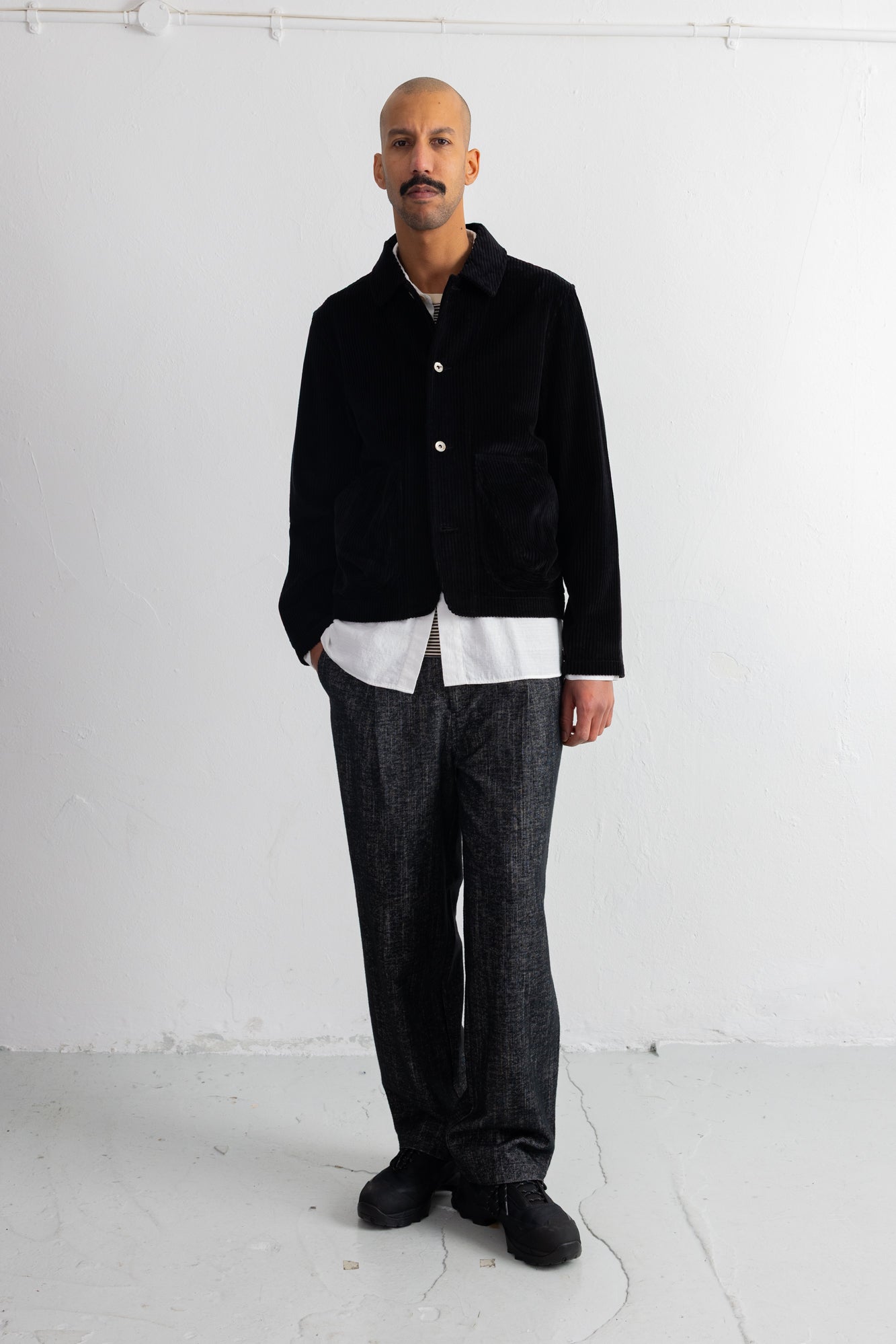 A man stands in a minimalist studio wearing a black corduroy jacket over a white shirt, paired with dark gray textured trousers and black shoes.