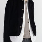 A man wearing a black corduroy jacket over a white button-up shirt and a beige-and-black striped shirt, paired with gray trousers, against a plain white background.