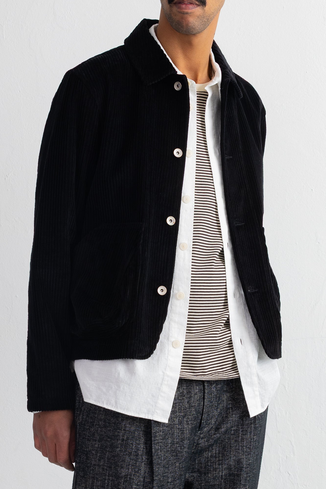 A man wearing a black corduroy jacket over a white button-up shirt and a beige-and-black striped shirt, paired with gray trousers, against a plain white background.