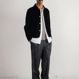 A man stands in a studio wearing a black corduroy jacket over a white shirt and gray textured trousers.