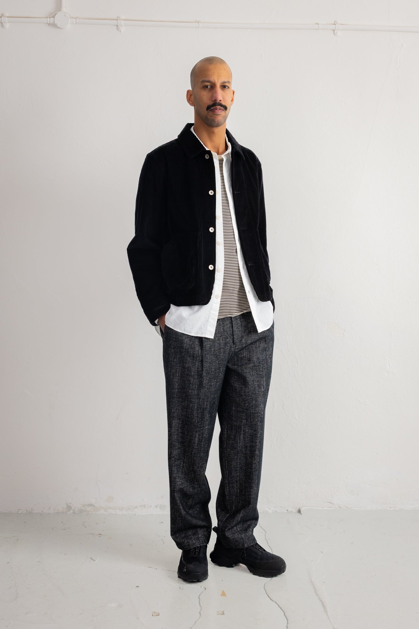 A man stands in a studio wearing a black corduroy jacket over a white shirt and gray textured trousers.
