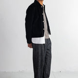 A man wearing a black corduroy jacket over a white t-shirt, paired with dark gray textured trousers and black shoes, standing against a white studio wall.