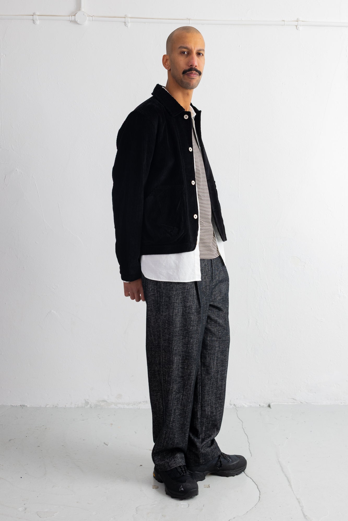 A man wearing a black corduroy jacket over a white t-shirt, paired with dark gray textured trousers and black shoes, standing against a white studio wall.