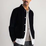 A man wearing a black corduroy jacket over a white shirt and striped sweater, paired with dark gray trousers, standing against a plain light gray studio background.