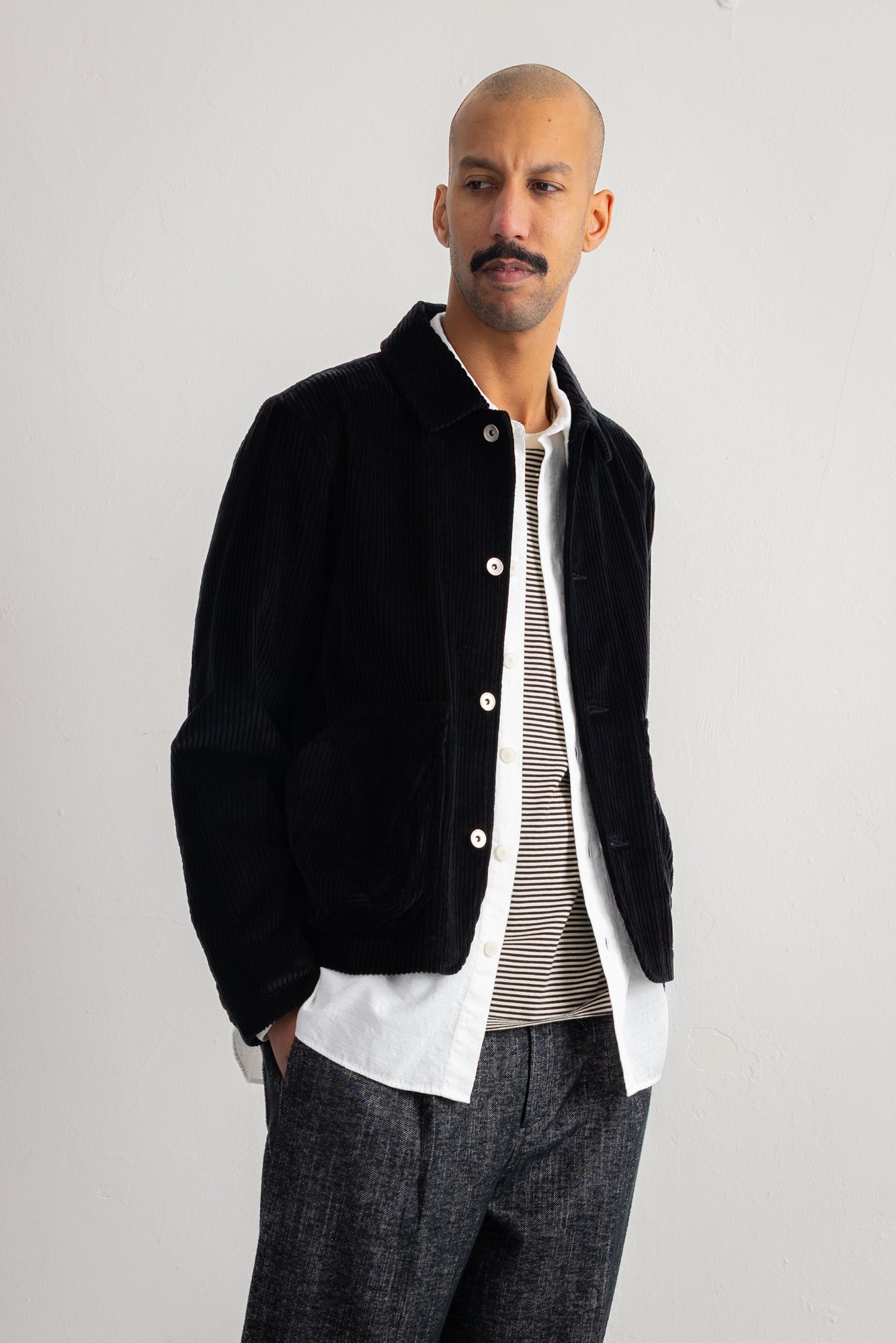 A man wearing a black corduroy jacket over a white shirt and striped sweater, paired with dark gray trousers, standing against a plain light gray studio background.