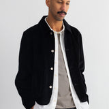 A man wearing a black corduroy jacket over a white shirt and a gray striped t-shirt, paired with dark gray trousers, standing against a white wall.