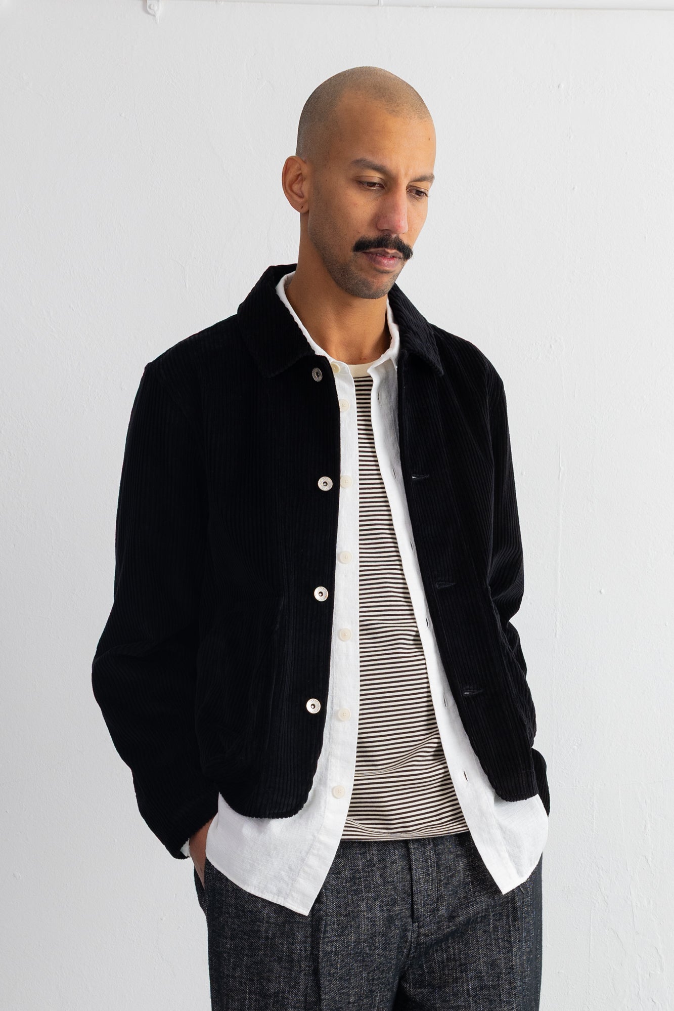 A man wearing a black corduroy jacket over a white shirt and a gray striped t-shirt, paired with dark gray trousers, standing against a white wall.