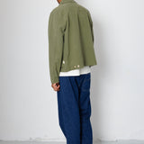 Person wearing a green jacket and blue jeans against a white wall