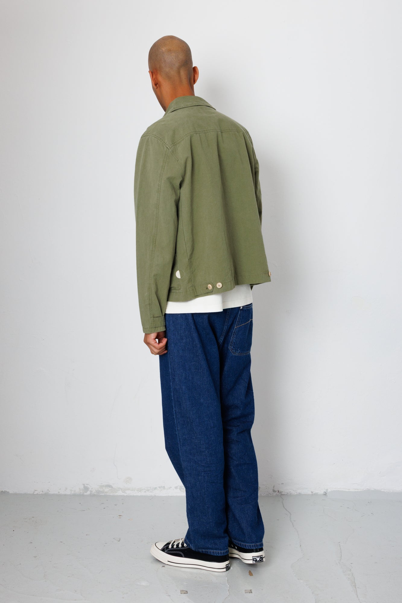 Person wearing a green jacket and blue jeans against a white wall