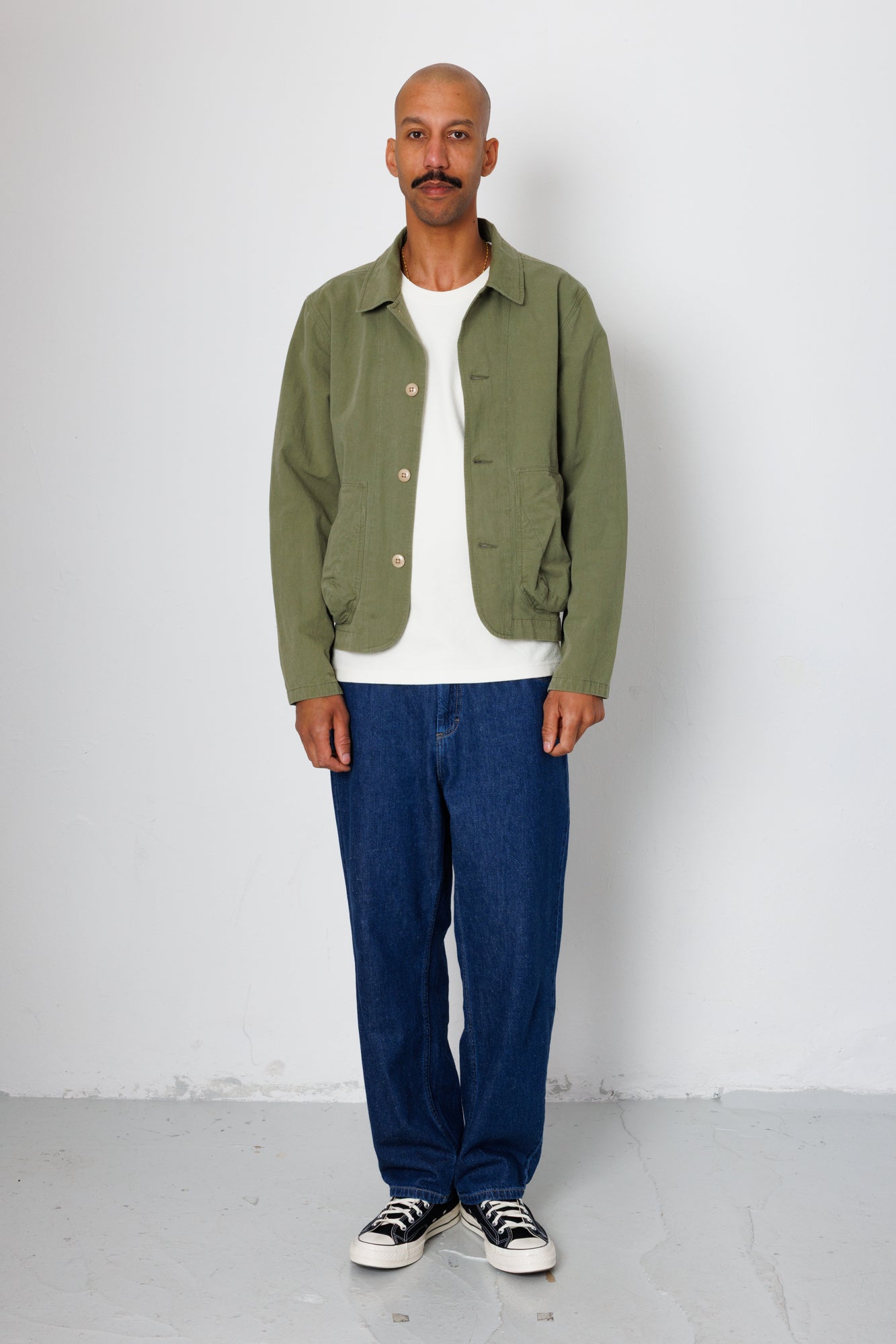 Man wearing a green jacket, white shirt, blue pants, and black sneakers against a white background
