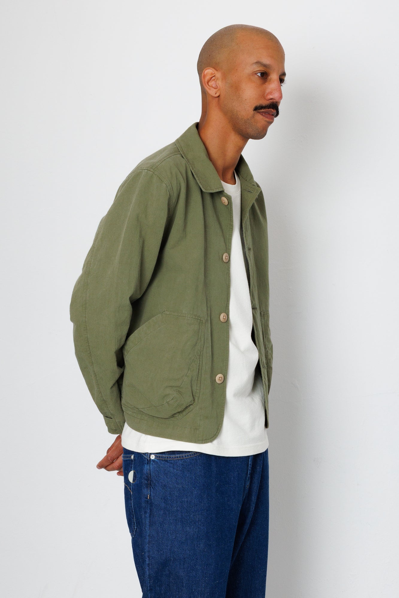 Man wearing a green jacket over a white shirt and blue jeans on a plain background