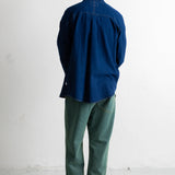 A man viewed from behind wearing a dark blue long-sleeve button-up shirt, green pants, and black-and-white sneakers, standing against a plain white wall.