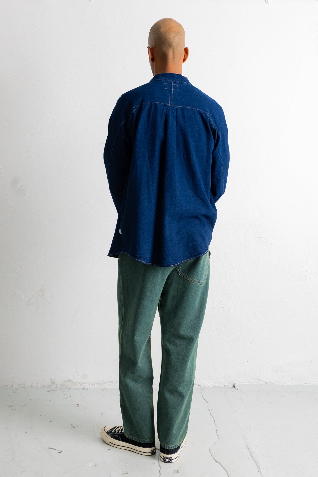 A man viewed from behind wearing a dark blue long-sleeve button-up shirt, green pants, and black-and-white sneakers, standing against a plain white wall.