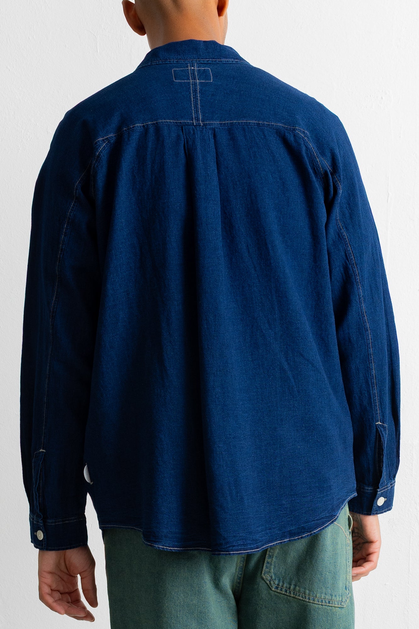 A person shown from the back wearing a dark indigo blue long-sleeve shirt and green pants against a plain white background.