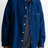 A person wearing a dark indigo blue denim button-up shirt jacket over a dark gray striped t-shirt and green jeans, shown from the neck down against a plain white background.