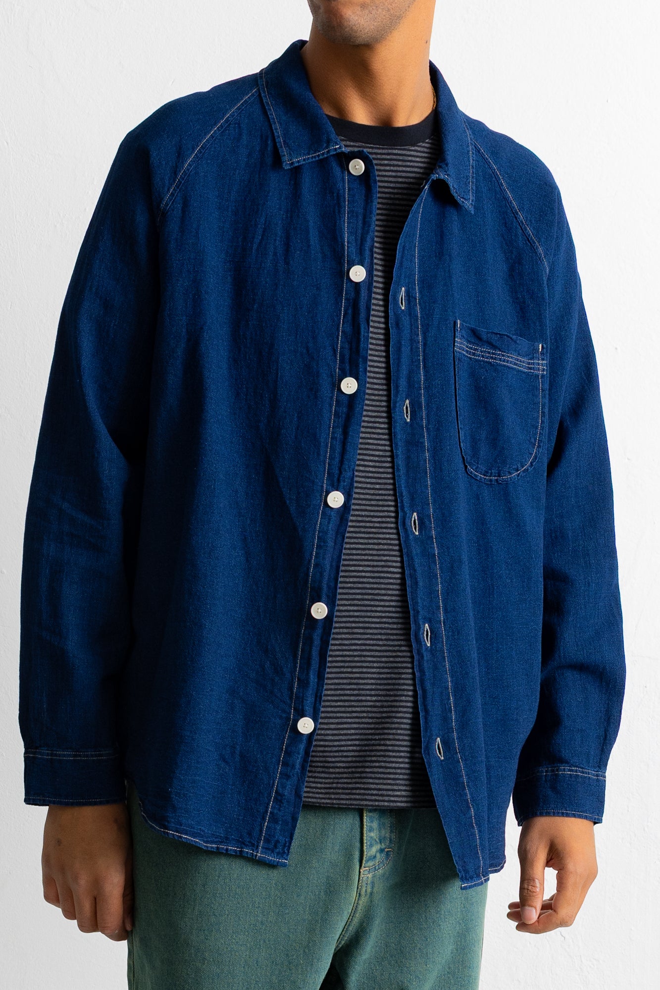 A person wearing a dark indigo blue denim button-up shirt jacket over a dark gray striped t-shirt and green jeans, shown from the neck down against a plain white background.