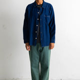 A man stands against a plain white wall wearing a dark blue button-up overshirt over a black t-shirt, paired with loose green pants and black-and-white sneakers.