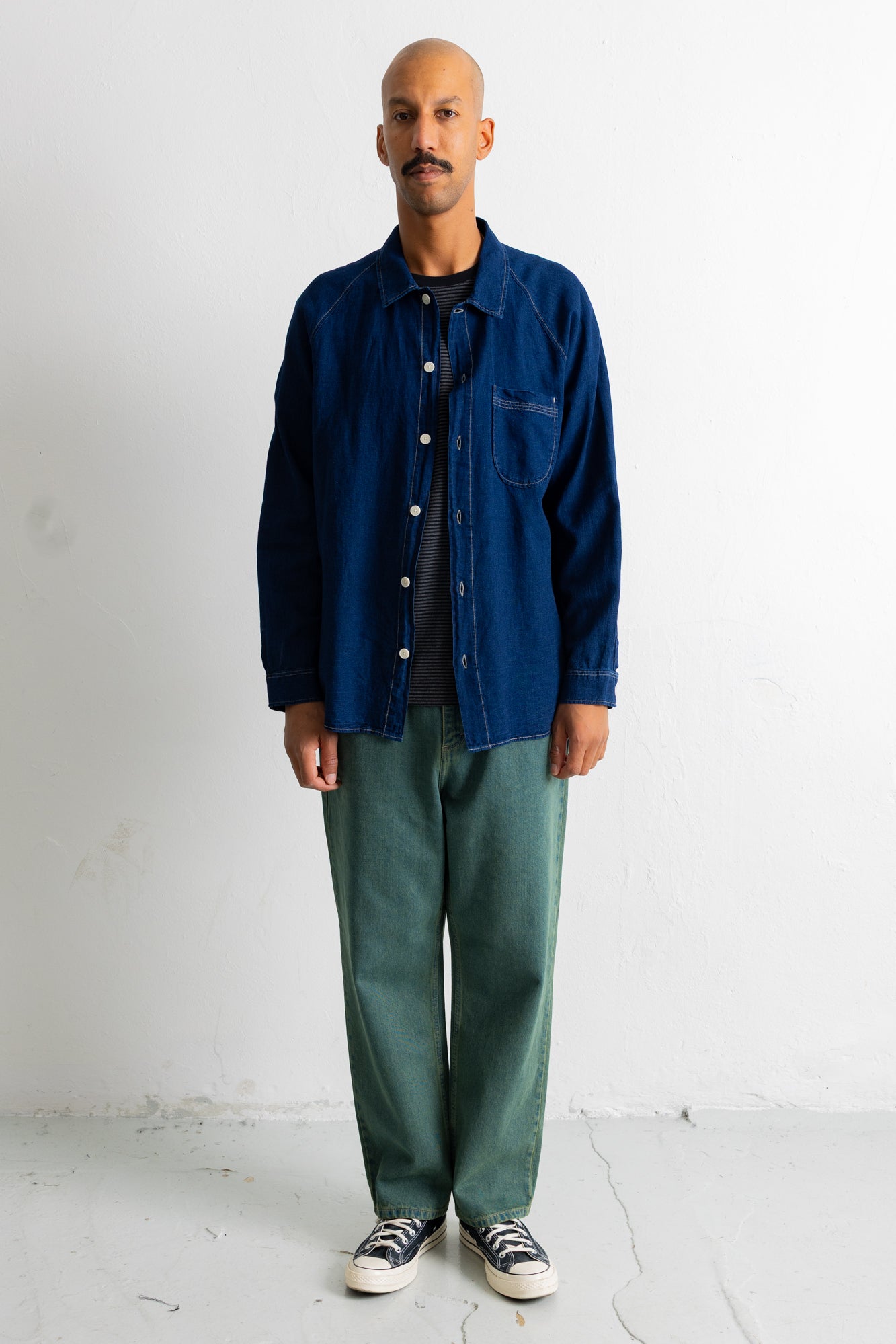 A man stands against a plain white wall wearing a dark blue button-up overshirt over a black t-shirt, paired with loose green pants and black-and-white sneakers.