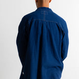 A man viewed from behind wearing a dark indigo blue long-sleeve button-up shirt and green pants, standing against a plain white wall.