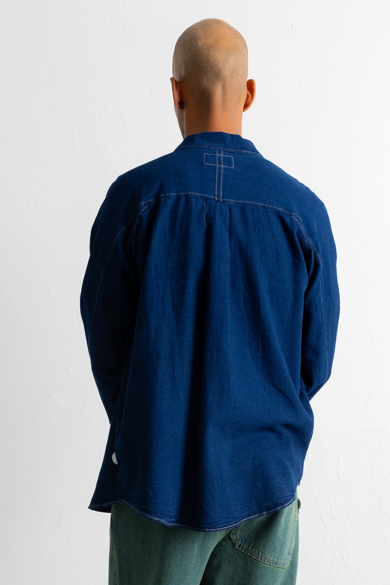 A man viewed from behind wearing a dark indigo blue long-sleeve button-up shirt and green pants, standing against a plain white wall.