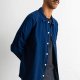A man wearing a dark indigo blue denim button-up shirt over a dark striped t-shirt and teal pants, photographed against a plain white wall.