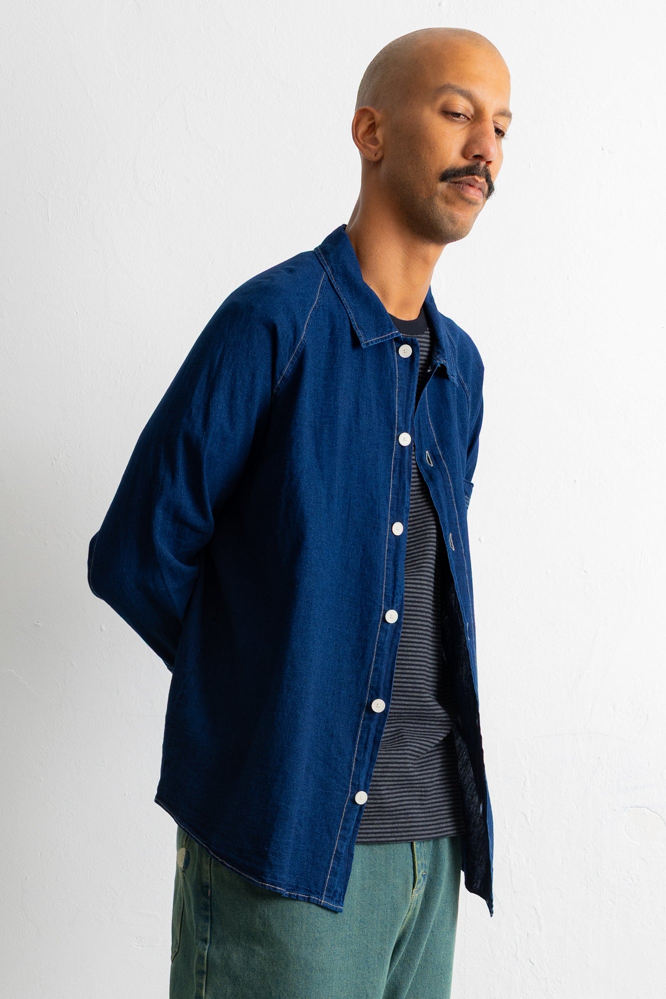 A man wearing a dark indigo blue denim button-up shirt over a dark striped t-shirt and teal pants, photographed against a plain white wall.