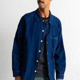 A man wearing a dark indigo blue denim work jacket over a dark striped shirt and teal pants, standing against a plain white wall.