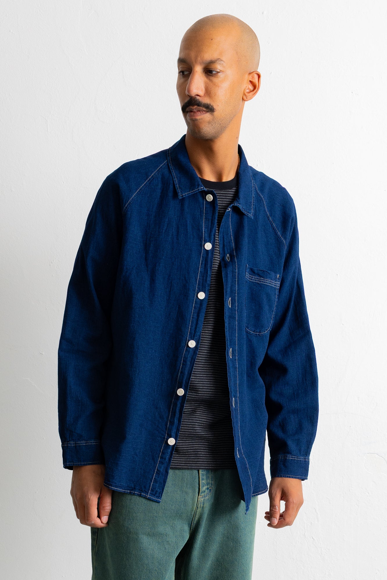 A man wearing a dark indigo blue denim work jacket over a dark striped shirt and teal pants, standing against a plain white wall.