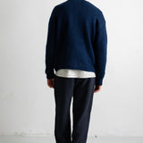 A man viewed from behind wearing a navy blue knit sweater over a white and navy striped shirt, dark navy pants, and black canvas sneakers, standing against a white wall.