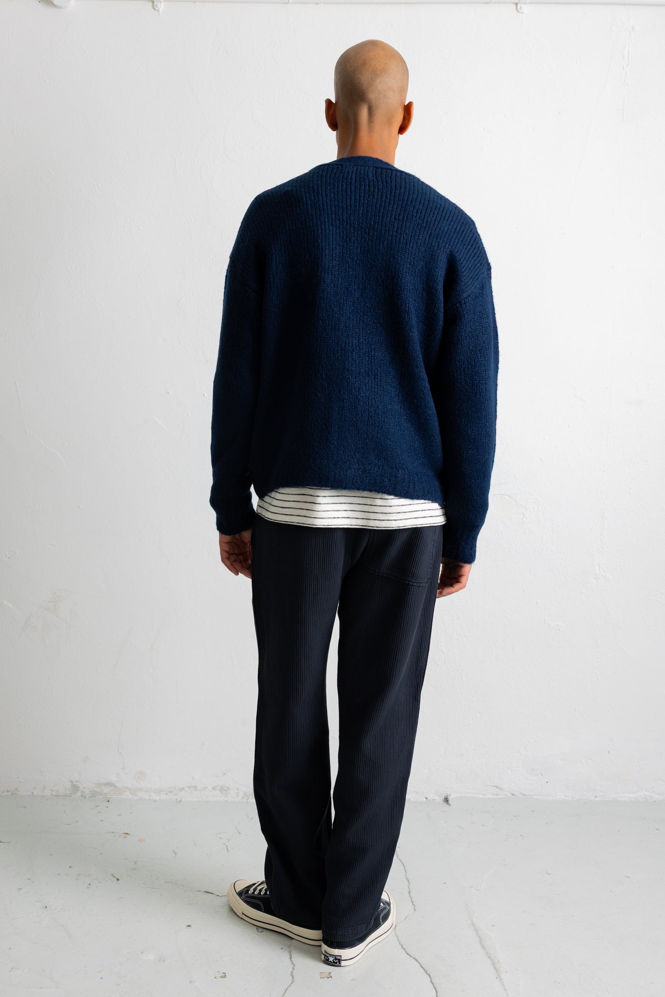 A man viewed from behind wearing a navy blue knit sweater over a white and navy striped shirt, dark navy pants, and black canvas sneakers, standing against a white wall.