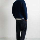A man viewed from behind wearing a dark navy blue knit sweater, dark navy trousers, and white sneakers, standing against a plain white wall.