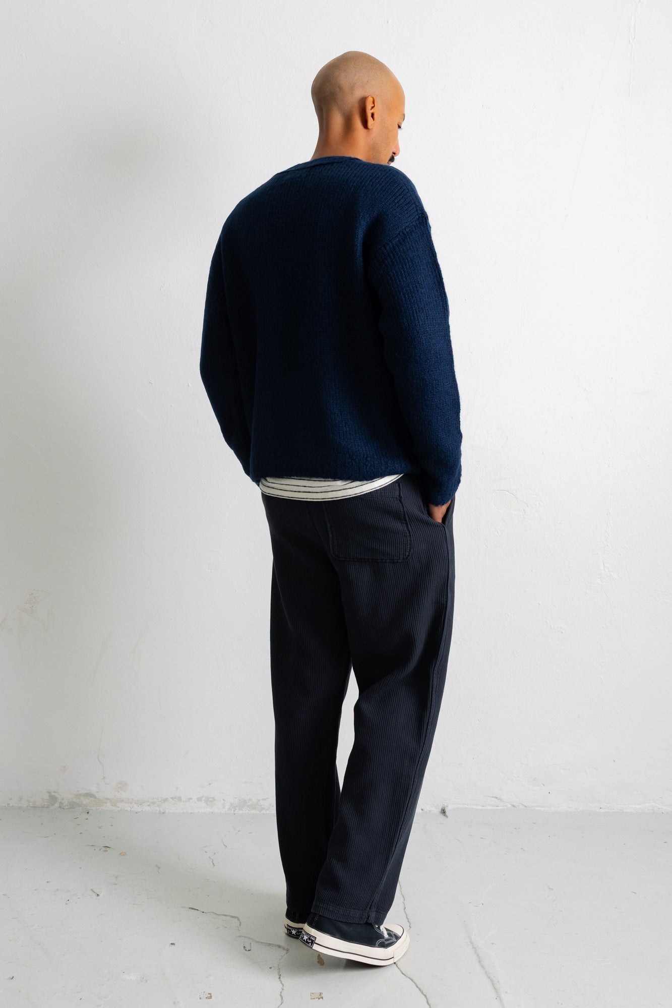 A man viewed from behind wearing a dark navy blue knit sweater, dark navy trousers, and white sneakers, standing against a plain white wall.