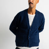 A man wearing a navy blue knit cardigan over a white and navy striped t-shirt, paired with dark trousers, standing against a white wall.