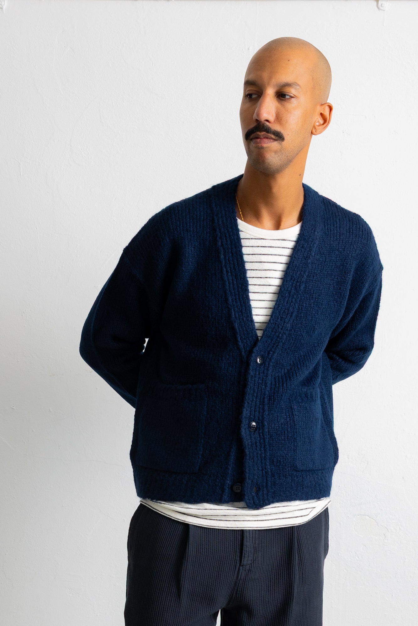 A man wearing a navy blue knit cardigan over a white and navy striped t-shirt, paired with dark trousers, standing against a white wall.