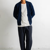 A man wearing a navy blue knit cardigan over a white and navy striped t-shirt, paired with navy blue sweatpants and black canvas sneakers, standing against a white wall.