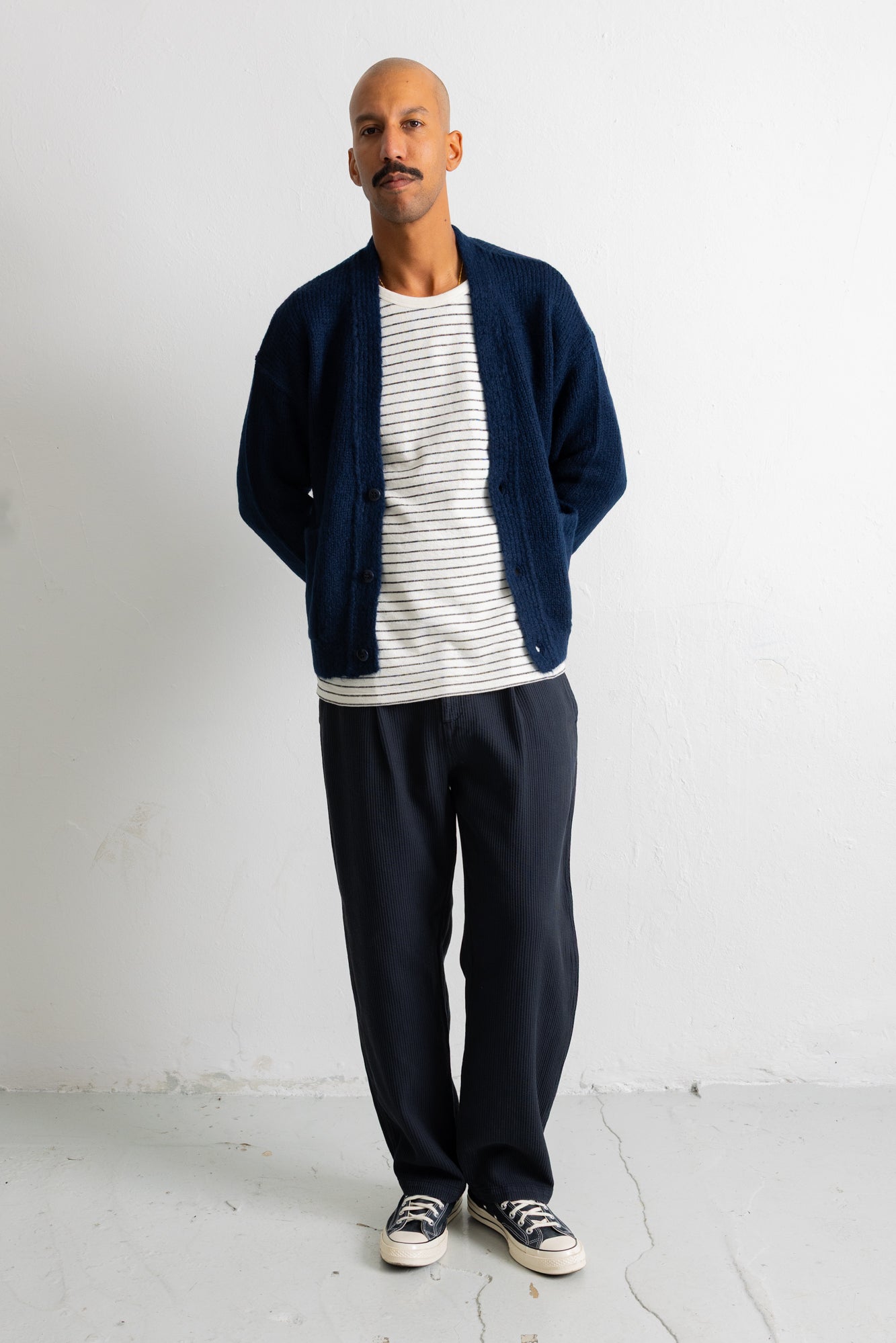 A man wearing a navy blue knit cardigan over a white and navy striped t-shirt, paired with navy blue sweatpants and black canvas sneakers, standing against a white wall.