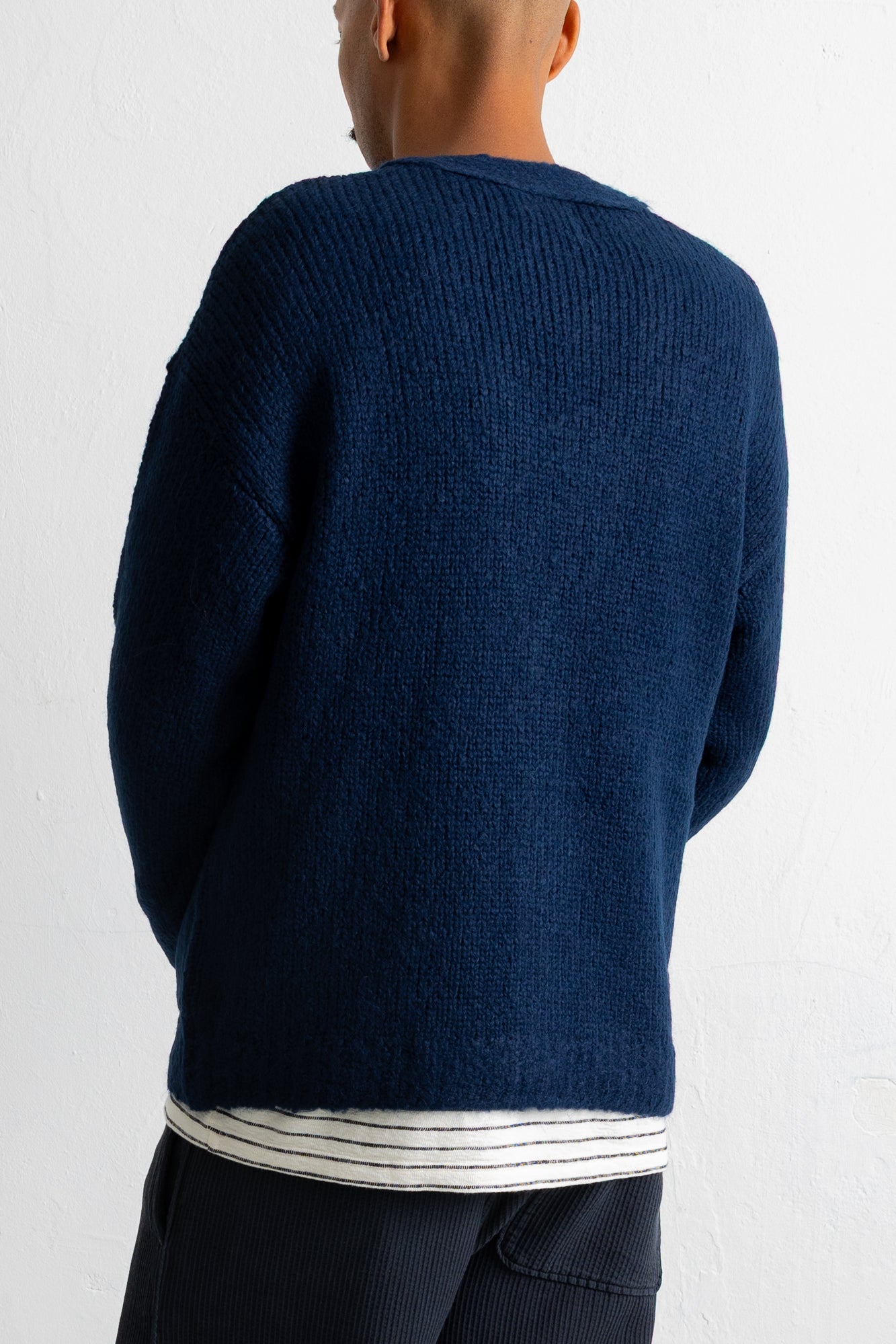 A person shown from the back wearing a dark navy ribbed knit sweater over a white striped shirt, against a plain white wall.