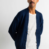 A man wearing a navy blue knit cardigan over a white and navy striped t-shirt, paired with dark trousers, standing against a plain white wall.