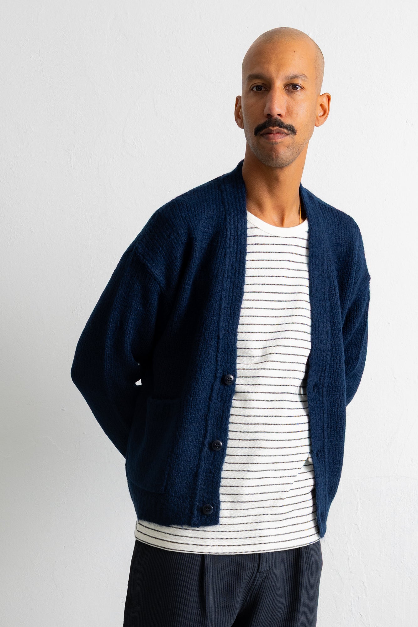 A man wearing a navy blue knit cardigan over a white and navy striped t-shirt, paired with dark trousers, standing against a plain white wall.