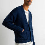 A man wearing a navy blue knit cardigan over a white and navy striped t-shirt, paired with dark trousers, standing against a light gray wall.