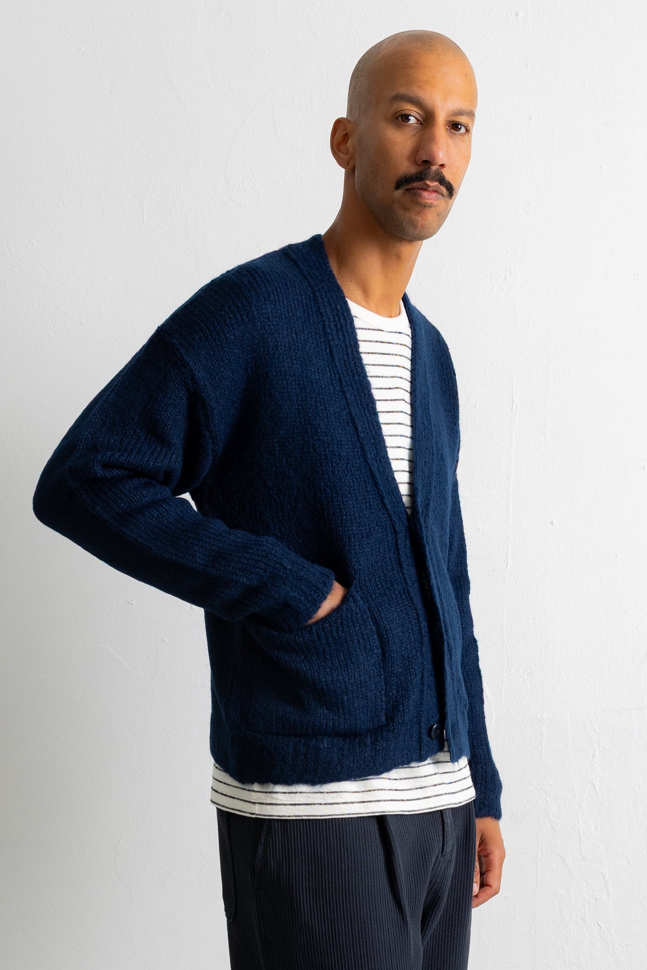 A man wearing a navy blue knit cardigan over a white and navy striped t-shirt, paired with dark trousers, standing against a light gray wall.