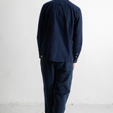 A man viewed from behind wearing a dark navy blue long-sleeve button-up shirt and matching navy blue trousers, standing against a plain white wall.