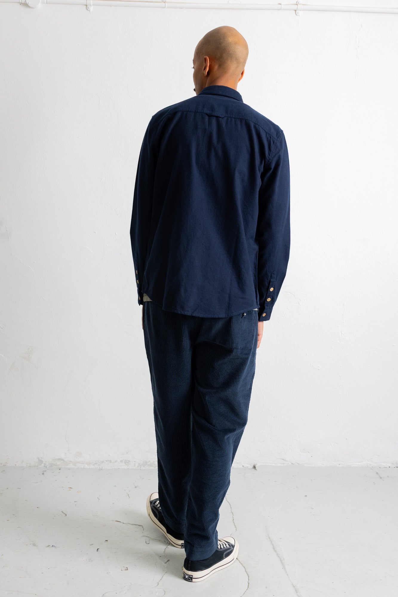 A man viewed from behind wearing a dark navy blue long-sleeve button-up shirt and matching navy blue trousers, standing against a plain white wall.