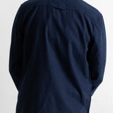 A man viewed from the back wearing a dark navy blue long-sleeve button-up shirt and matching navy pants against a plain white background.