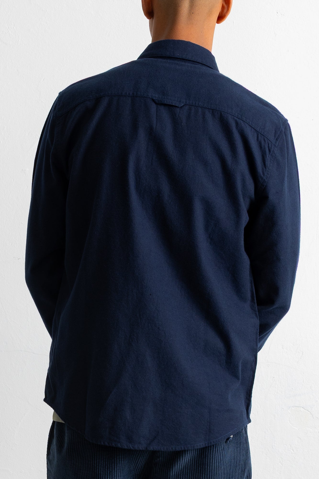 A man viewed from the back wearing a dark navy blue long-sleeve button-up shirt and matching navy pants against a plain white background.