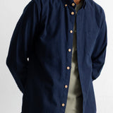 A man wearing a navy blue long-sleeve button-down shirt with a chest pocket, layered over a light olive green t-shirt, against a plain white background.